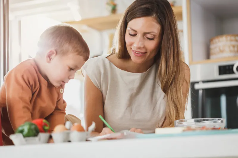 A parent sitting beside a young child, helping with an activity at the kitchen table
